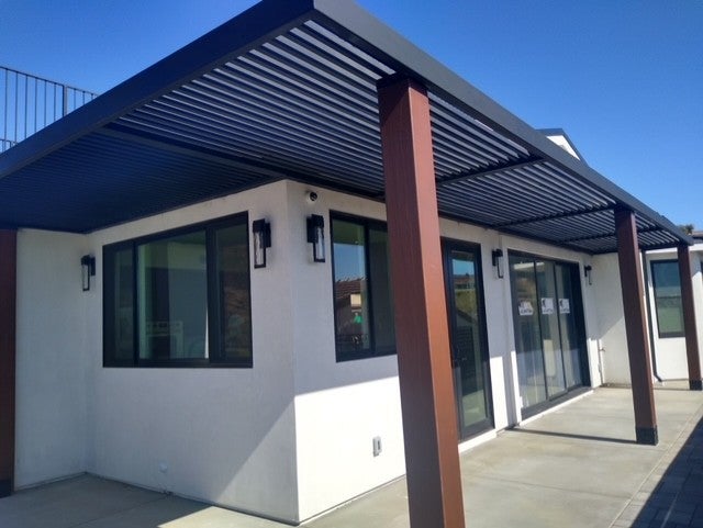 White house with black pergola and brown posts over a patio.