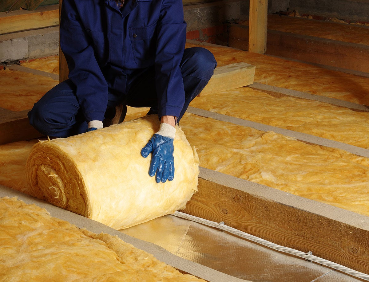 Worker installing yellow fiberglass insulation in attic rafters.